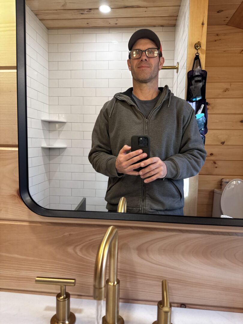 Man wearing glasses taking a selfie in a bathroom mirror.